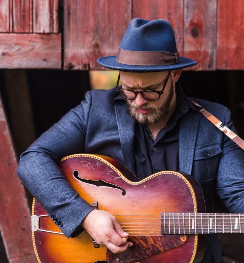 man-holding-jazz-guitar-standing-beside-brown-wooden-wall-1551018